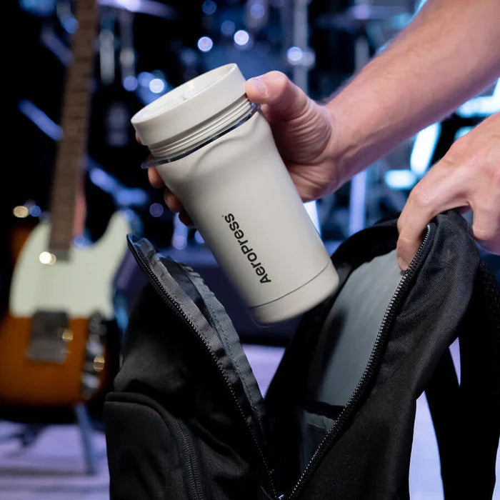 Person holding a white Aeropress coffee maker with a blurred background of musical instruments.