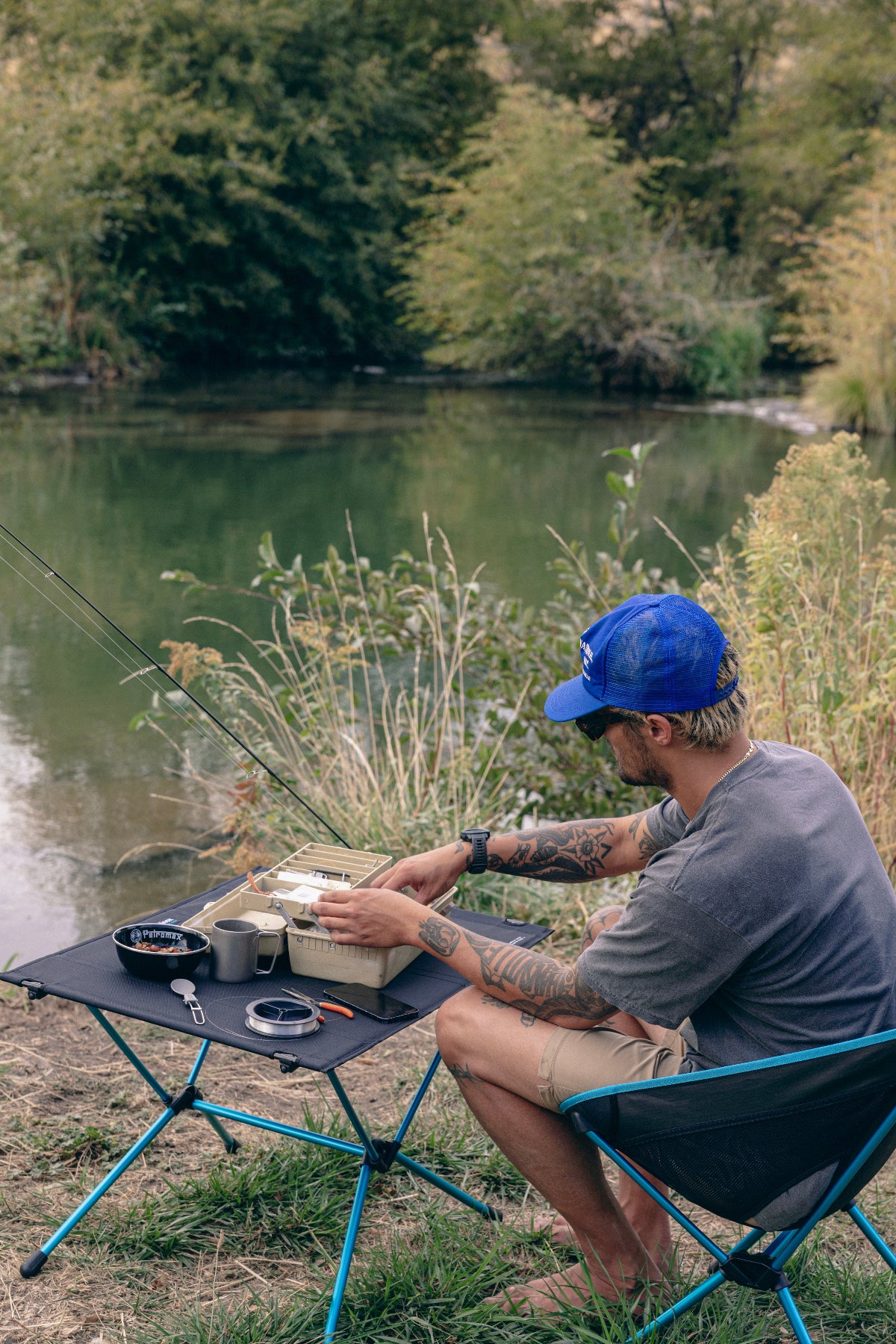 Man sitting by a lake with a portable stove and cooking equipment, wearing a blue cap sitting on Helinox camp furniture