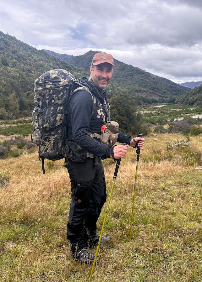 Man with a large backpack and hiking poles standing in a grassy field with mountains in the background hiking in New Zealand