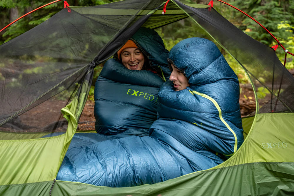 Two people in blue sleeping bags hiking inside a green tent with a forest background