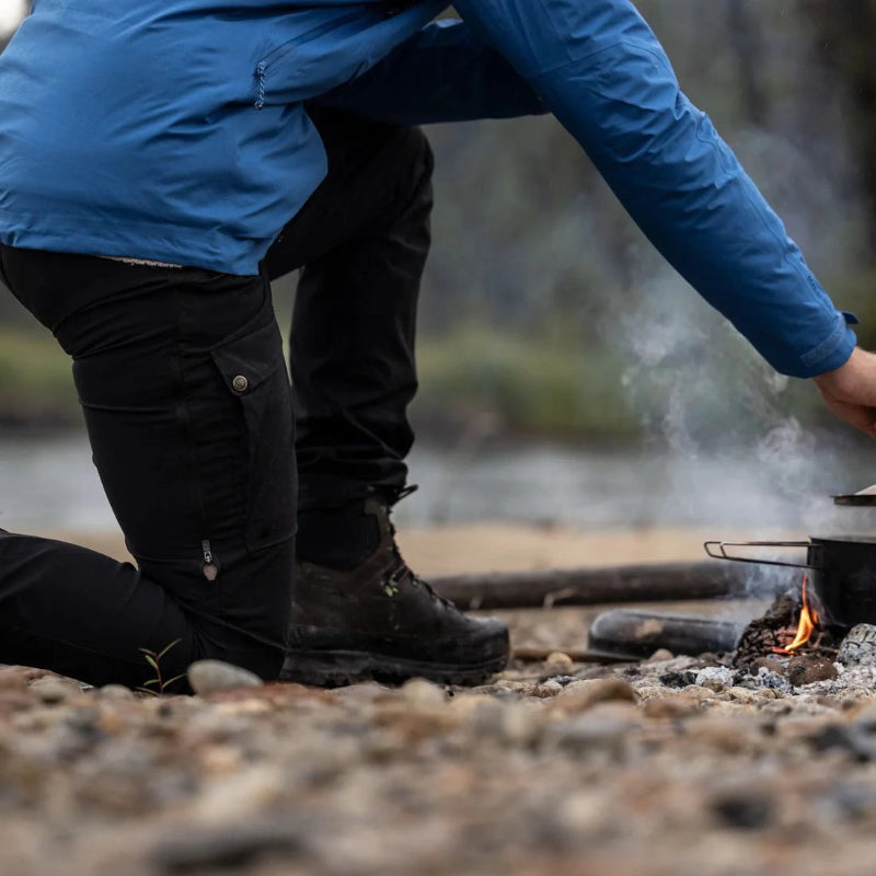 Person in blue jacket and black pants cooking over a campfire by a body of water.