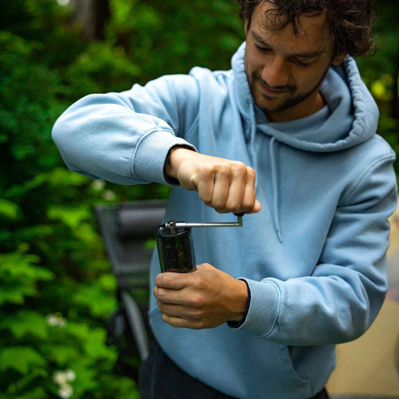 Man in light blue hoodie using a manual coffee grinder outdoors