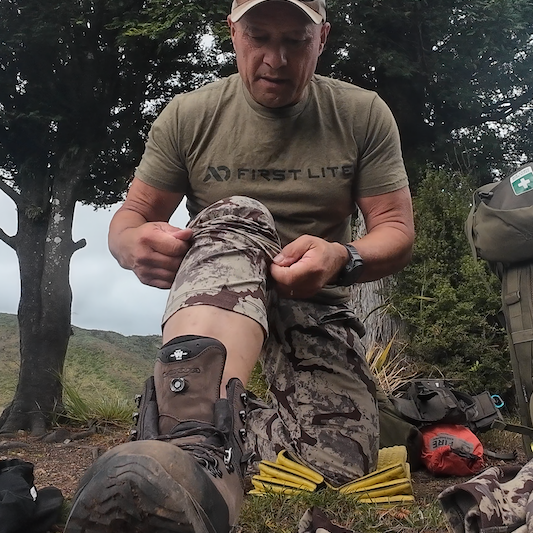 Man in outdoor setting wearing a 'First Lite' shirt and camouflage pants, sitting on the ground with trees in the background.