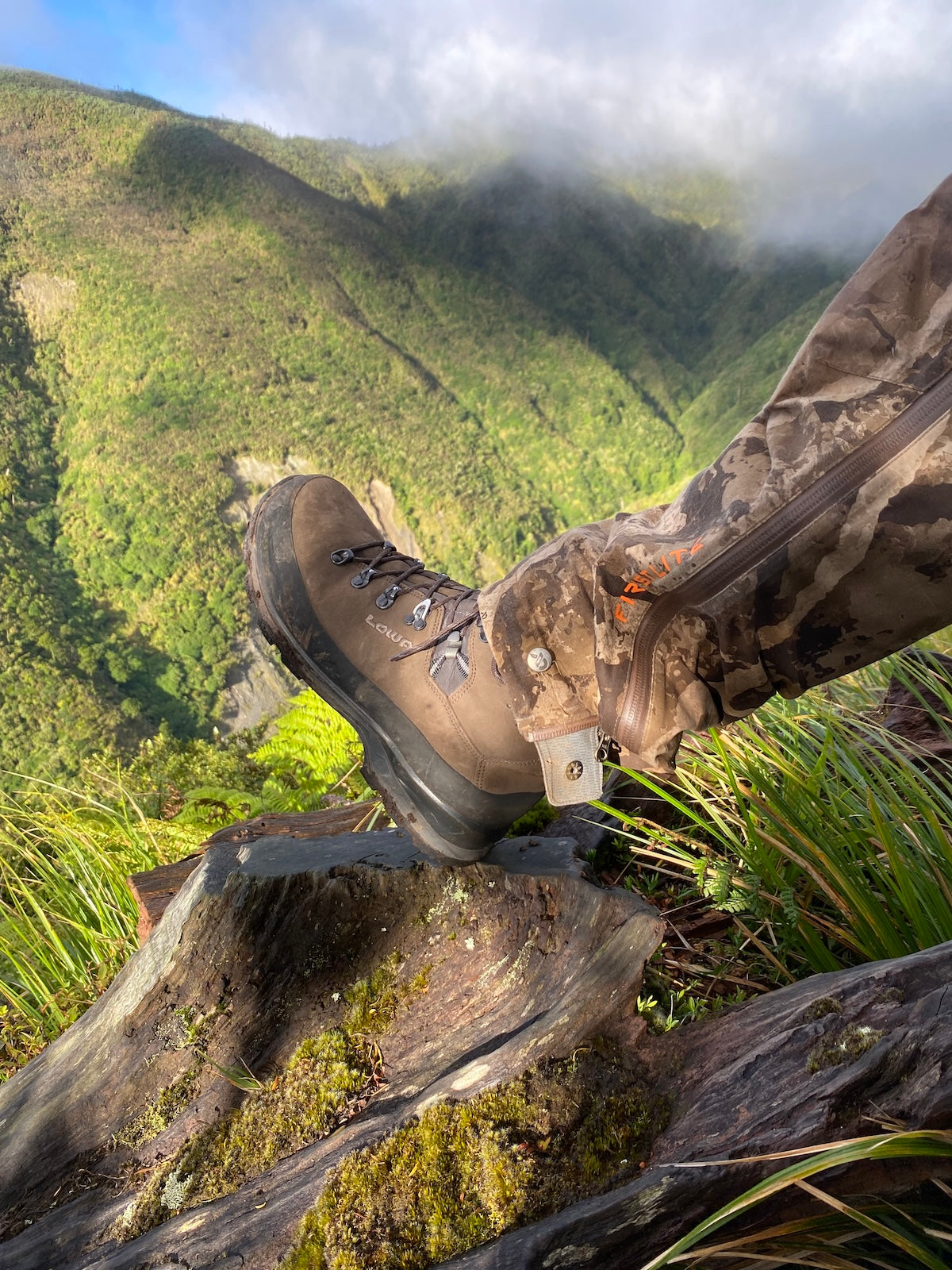 Male wearing Lowa hiking boots on a mountain in New Zealand with greenery and clouds in the background