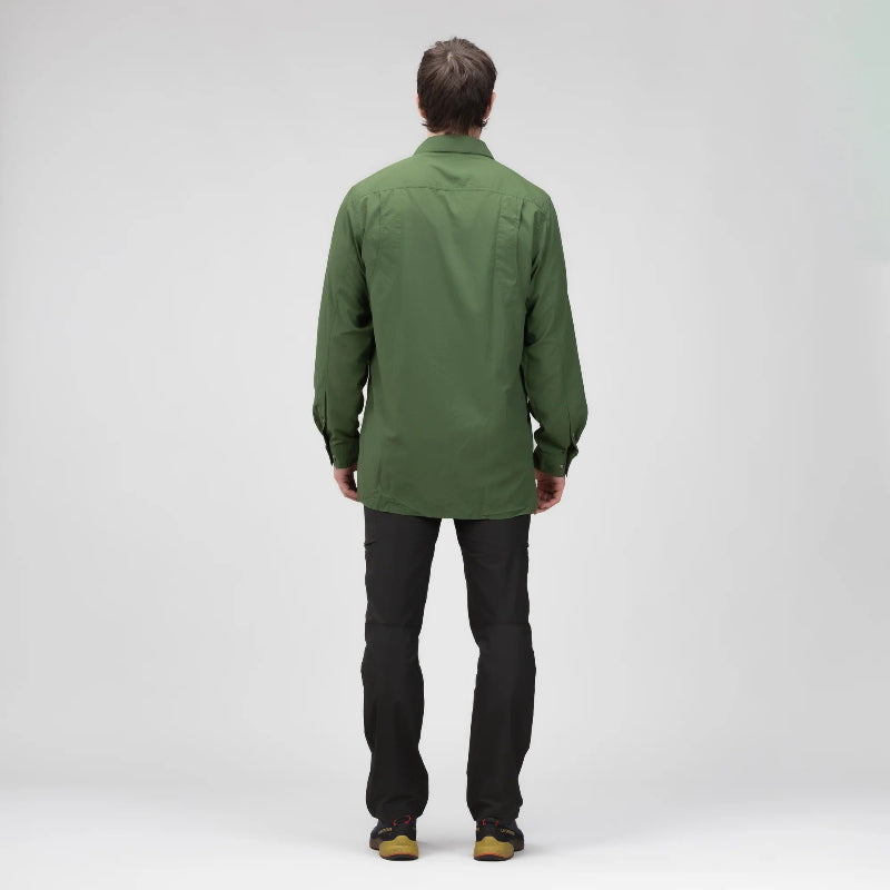 A man viewed from behind wearing an olive green long-sleeve shirt and black pants, standing against a light gray studio background.