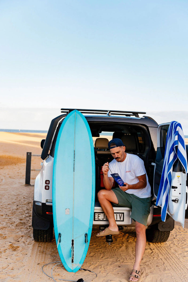 Man sitting in an open car door with a surfboard on a beach