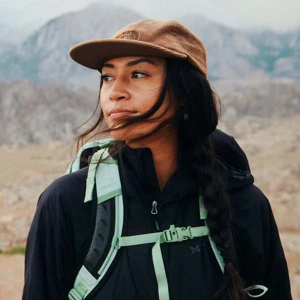 Person wearing a brown cap and black jacket with a green backpack in a mountainous landscape
