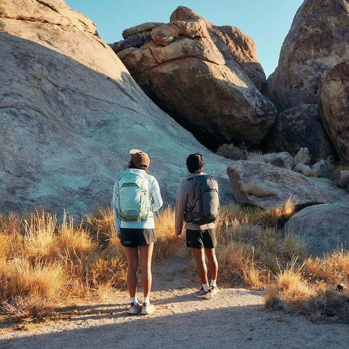 Two hikers with backpacks walking on a trail through rocky terrain.