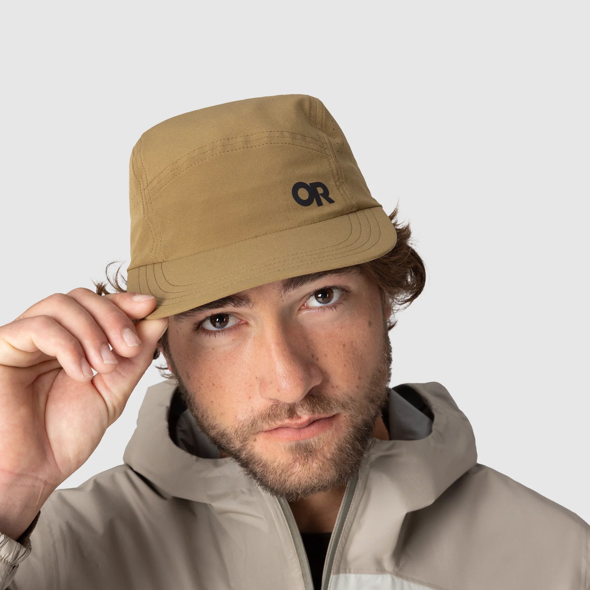 Man wearing a beige Outdoor hat with a logo, holding it with one hand, on a light gray background.
