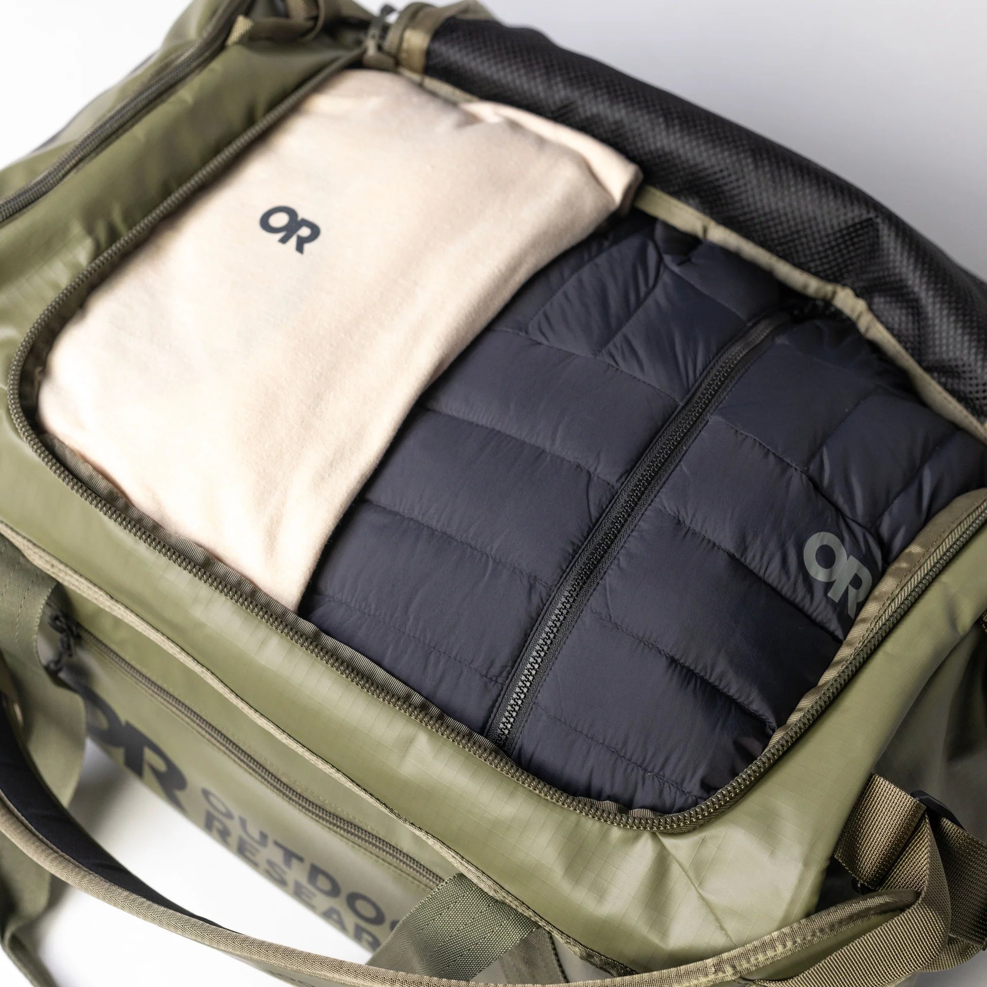 Green backpack with a black quilted jacket and beige item inside, featuring the 'OR' logo.