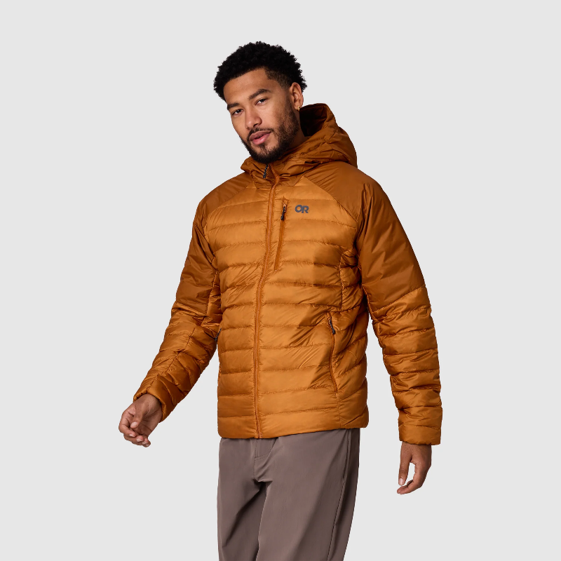 A man wearing a hooded quilted puffer jacket in a warm camel-brown color, paired with gray pants, photographed against a light gray studio background.