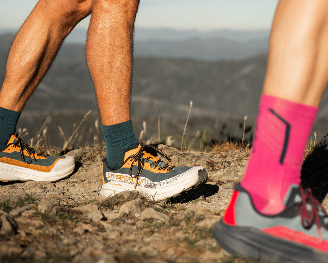 Close-up outdoor shot of two hikers' legs and feet on a rocky trail, wearing trail running shoes, dark teal socks, and bright pink socks with black graphics. The background shows blurred mountains under a pale sky.