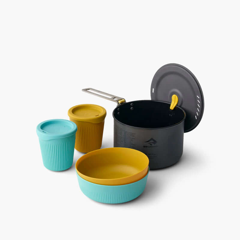 A set of camping cookware including a black hard-anodised aluminium pot with a yellow lid, and blue and yellow Passage dinnerware set which includes bowls and cups.