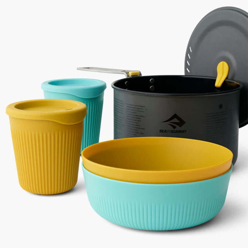 A set of camping cookware including a black hard-anodised aluminium pot with a yellow lid, and blue and yellow Passage dinnerware set which includes bowls and cups.