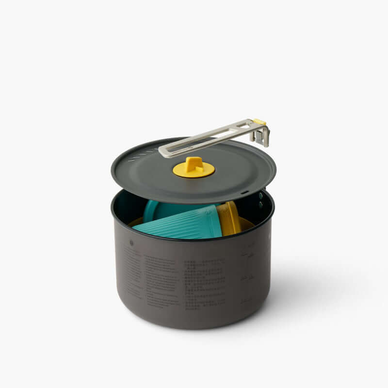 A set of camping cookware including a black hard-anodised aluminium pot with a yellow lid, and blue and yellow Passage dinnerware set which includes bowls and cups. All stacked inside the pot