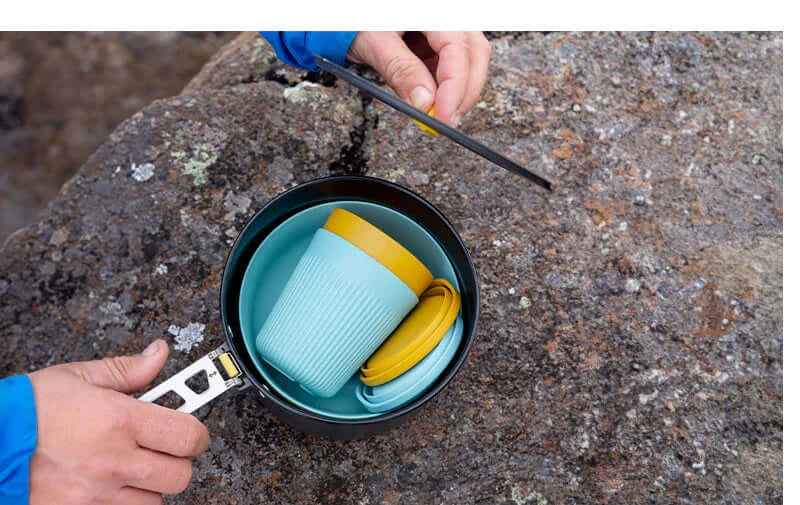 A set of camping cookware including a black hard-anodised aluminium pot with a yellow lid, and blue and yellow Passage dinnerware set which includes bowls and cups.