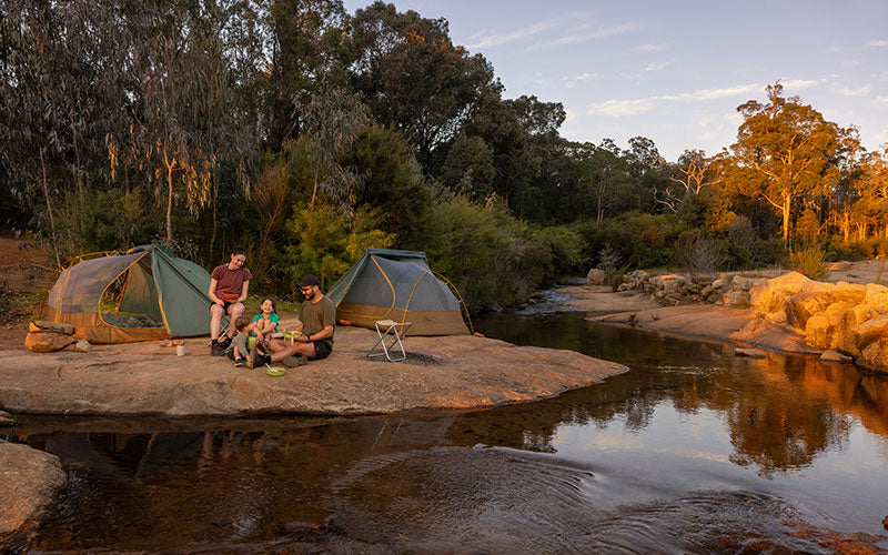 Two tents set up by a river with people sitting around, surrounded by trees.