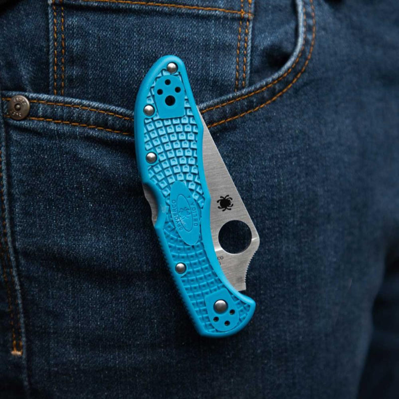 Blue folding knife with textured handle and metallic blade peeking out from a blue denim pocket.