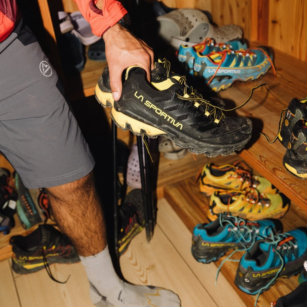 A person in athletic wear holds up a black La Sportiva trail running shoe with yellow accents, with other shoes and ski boots visible on wooden shelves in the background.