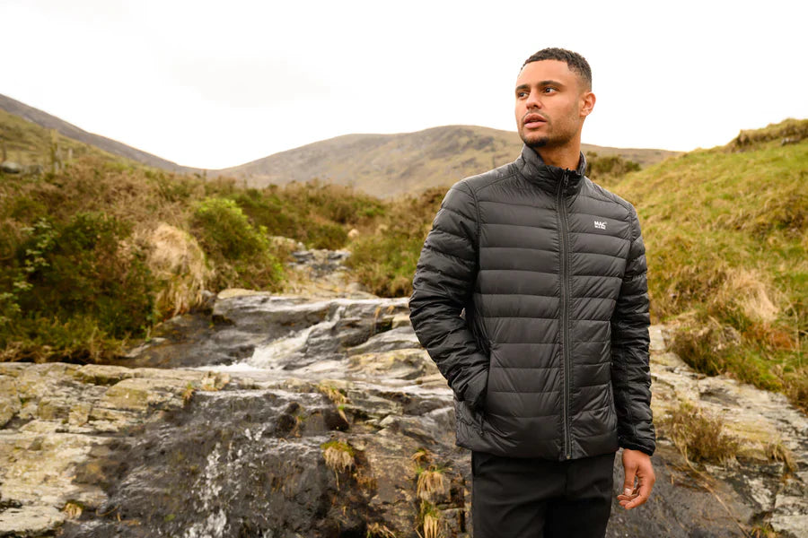 Man wearing a black puffer jacket standing in a mountainous landscape with a stream.