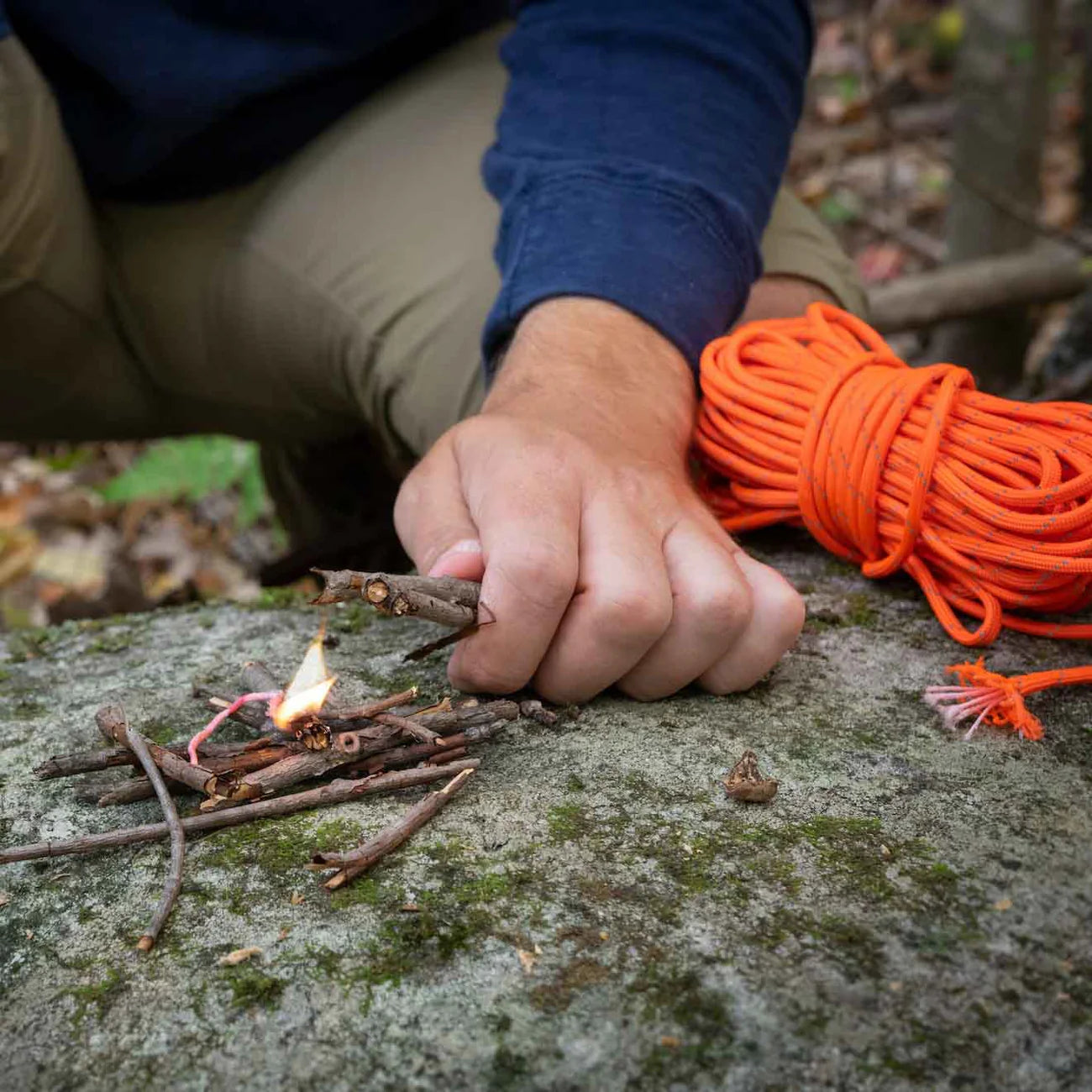 Person starting a fire with sticks, tinder cord and a lighter outdoors, surrounded by orange rope.
