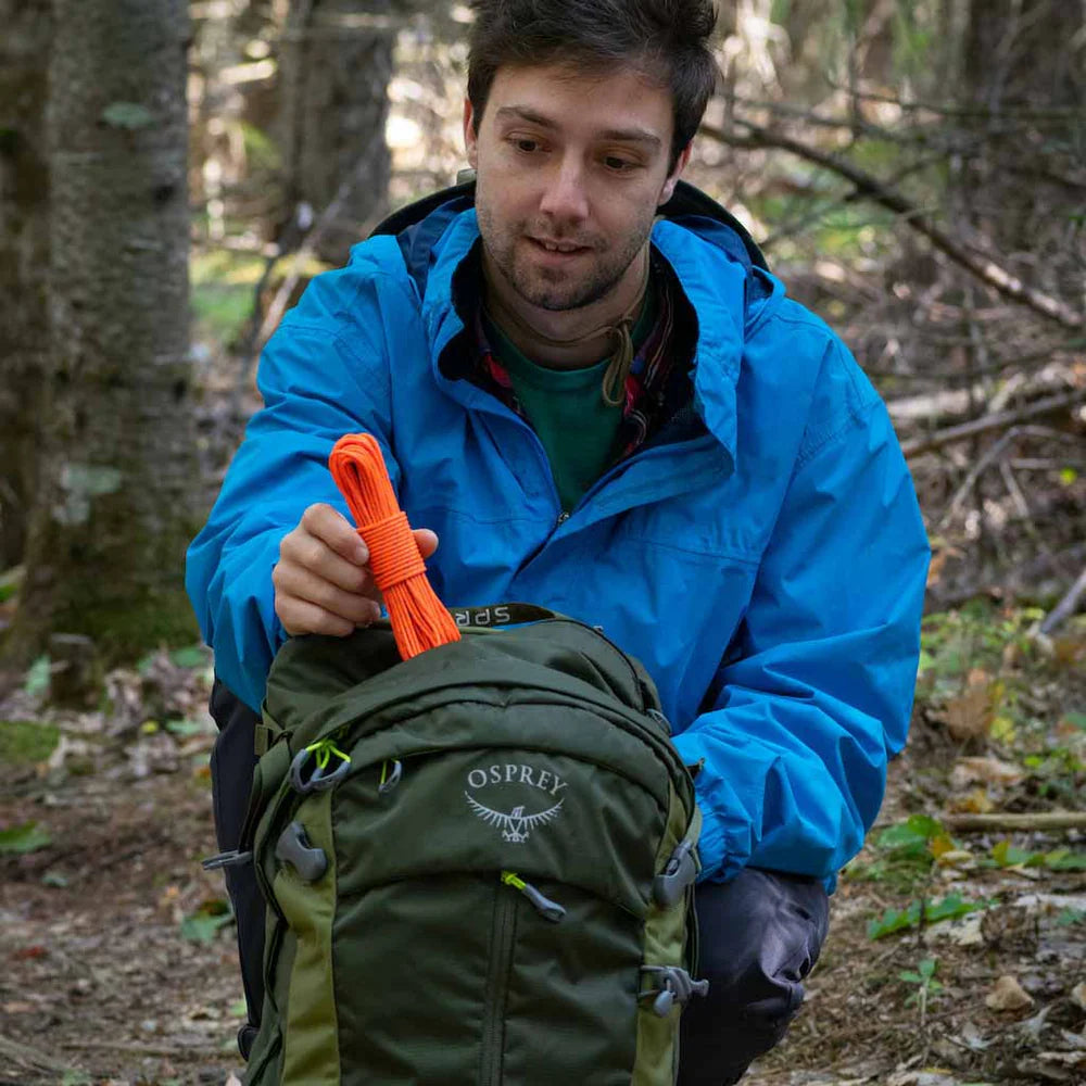 Man in blue jacket holding orange rope and green Osprey backpack in forest