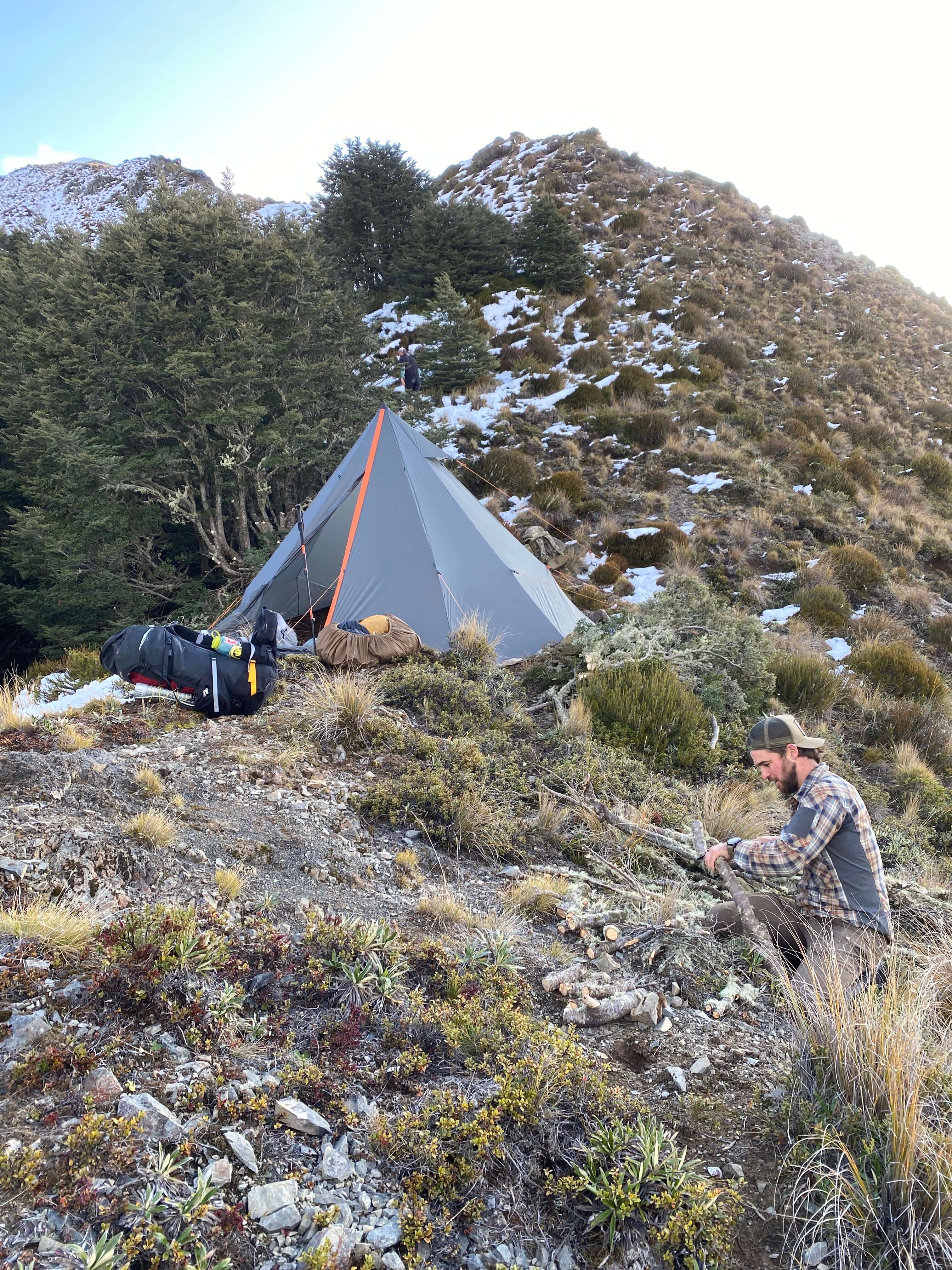 A person standing next to an Ultralight 4-6 Person Tipi Tent in a natural outdoor setting with trees and mountains in the background.