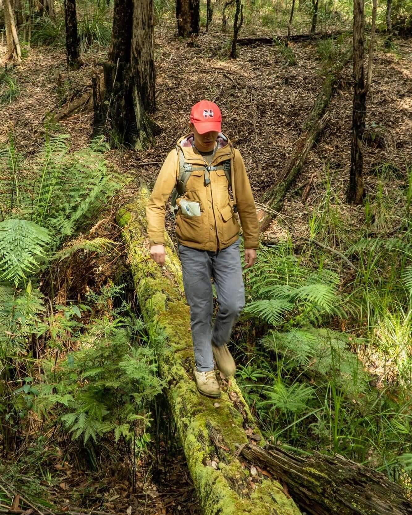 Instructor standing on a log in a forested area, wearing a yellow jacket and a red cap, conducting a outdoor navigation course.