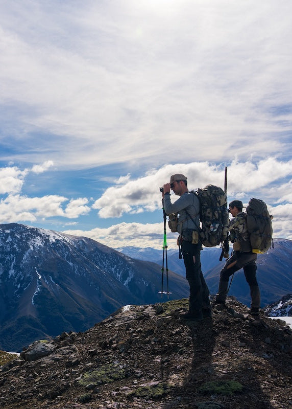 Two hikers with backpacks on a mountain peak hiking new zealand under a blue sky with clouds.
