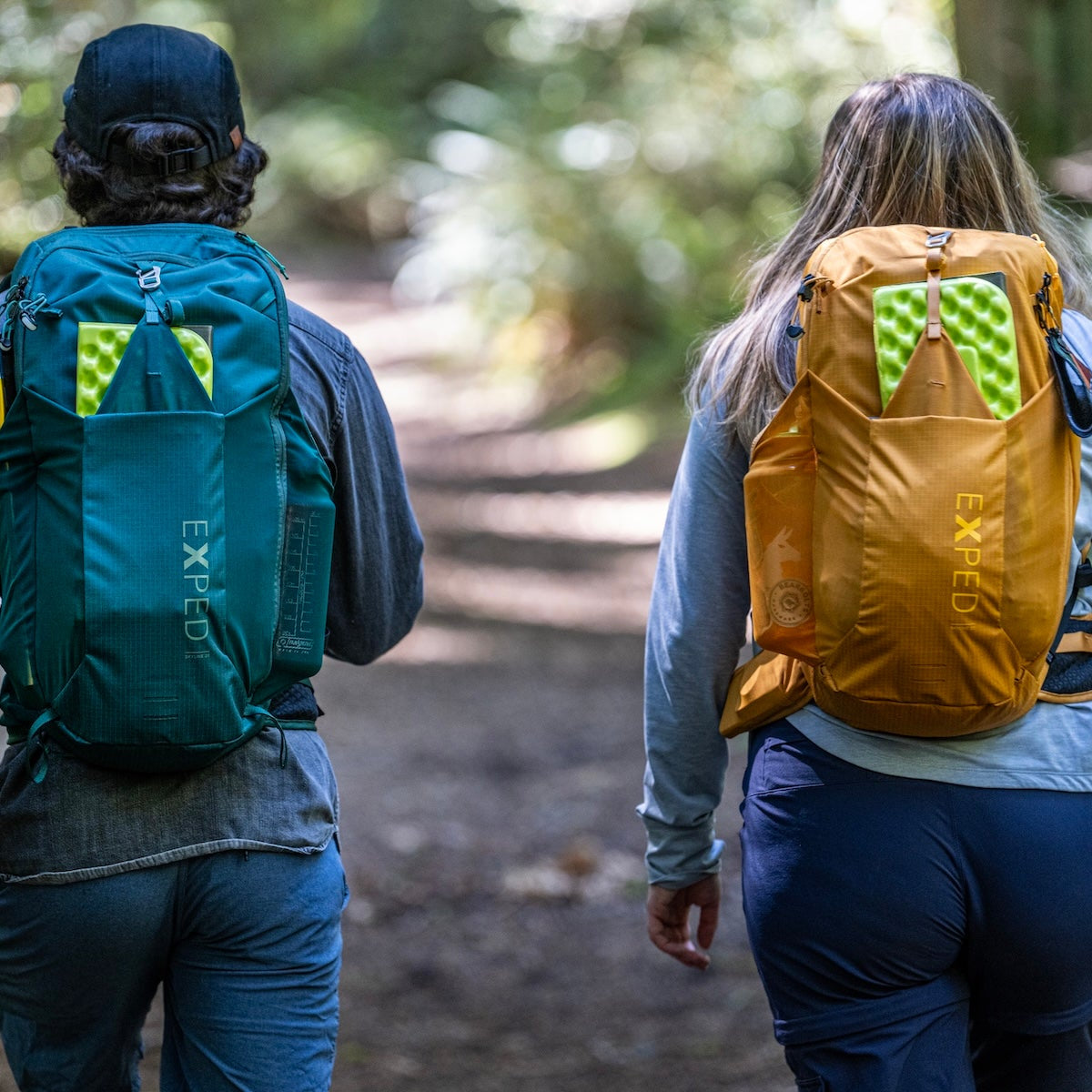 Two hikers viewed from behind on a forest trail, each wearing an Expedition backpack. The hiker on the left wears a teal backpack, and the hiker on the right wears a mustard yellow backpack, both with bright green reflective accents on the upper back.