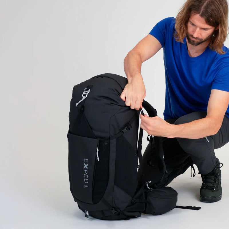 Person adjusting a black backpack with 'Exped' branding on a light gray background