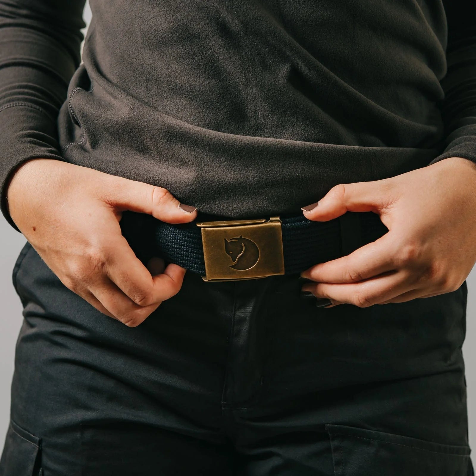 Person wearing a black belt with a brass buckle, adjusting it on a gray background