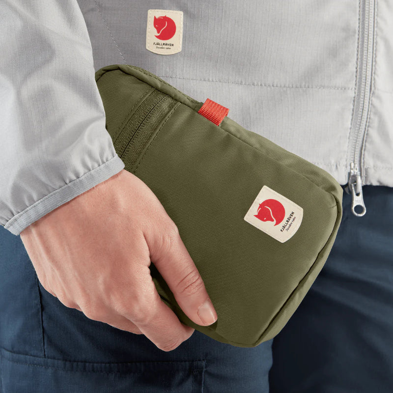 Person holding a green pouch with a brand logo, wearing a light gray jacket and dark pants.