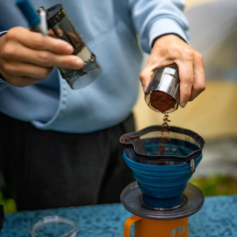 Person pouring coffee grounds into a blue coffee filter over an orange mug.