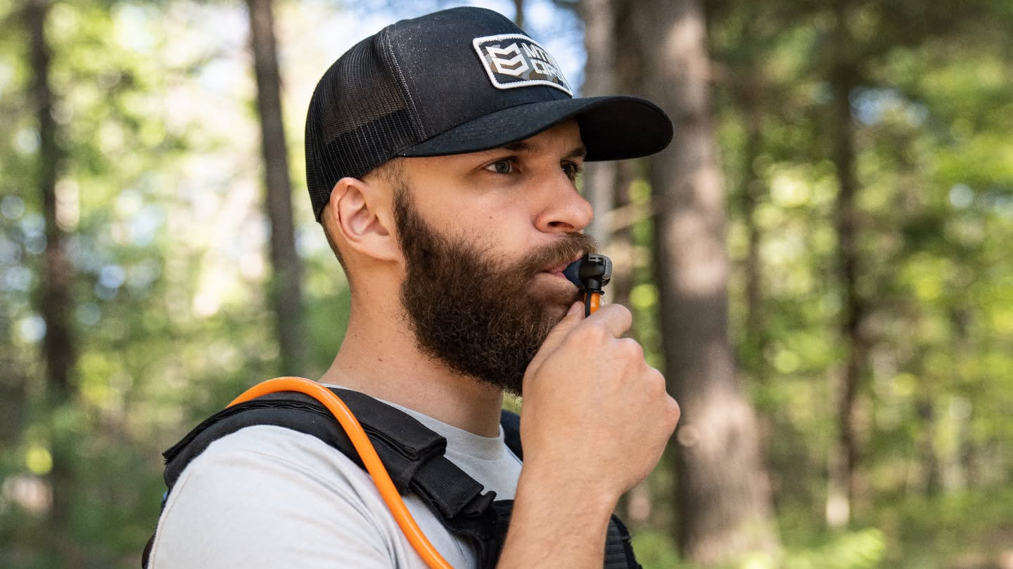 A bearded man wearing a black trucker hat with a white logo and a hydration backpack in a forest setting, drinking from an orange-capped water bottle.