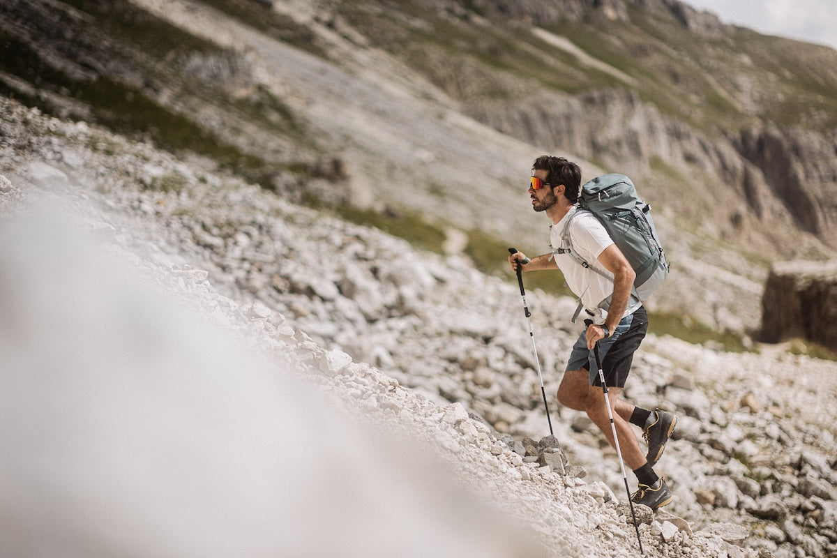 A man hiking up a rocky mountain slope using trekking poles and wearing a large gray backpack, with a blurred natural mountain background.