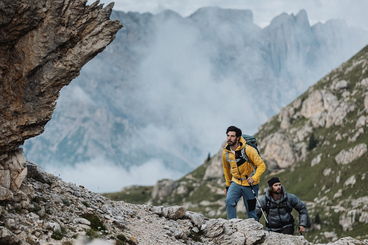 Two male hikers with backpacks traverse a rocky mountain path, with misty peaks and steep slopes in the background.