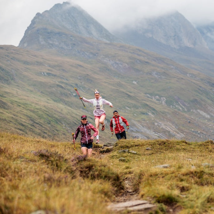 Three trail runners are seen from behind running up a grassy mountain path with rugged peaks and clouds in the background.