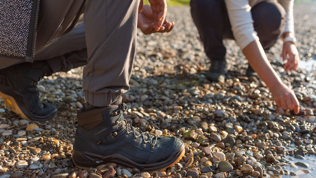 Two people crouching on a pebbly beach, one person is wearing boots.