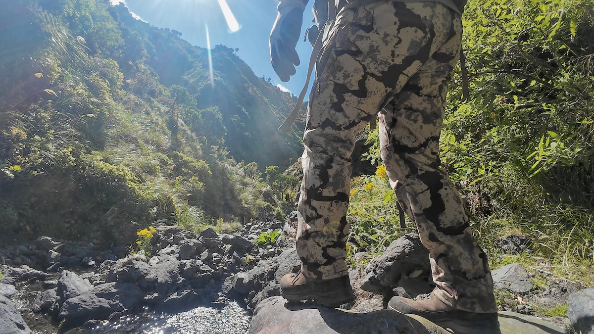 Person wearing camouflage pants standing on a rocky trail with mountains in the background