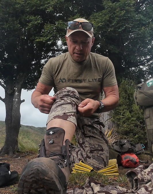 Man in outdoor setting wearing a 'First Lite' shirt and camouflage pants, sitting on the ground with trees in the background.