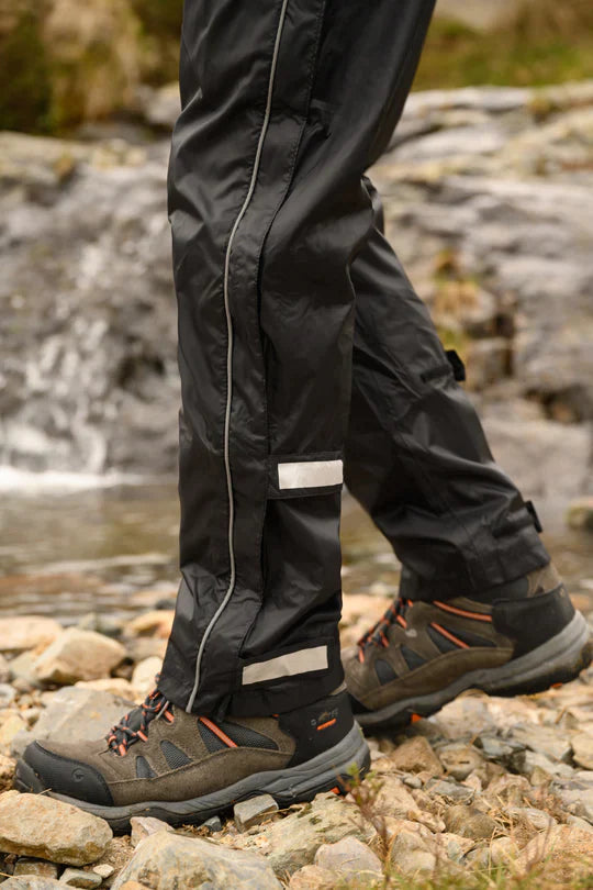 A man wearing packable rain overpants from the waist down, standing on a riverbed