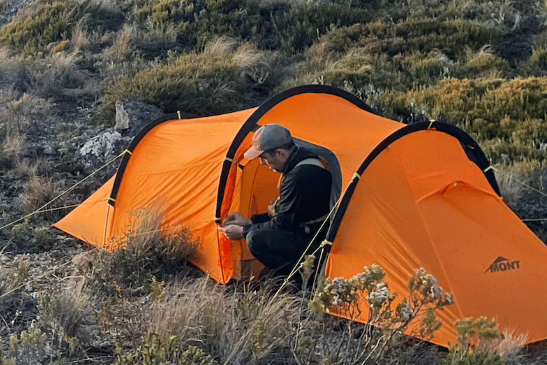 An orange dome camping tent pitched on a grassy, rocky hillside, with a person sitting at the entrance. The background shows natural outdoor terrain in muted brown and green tones.