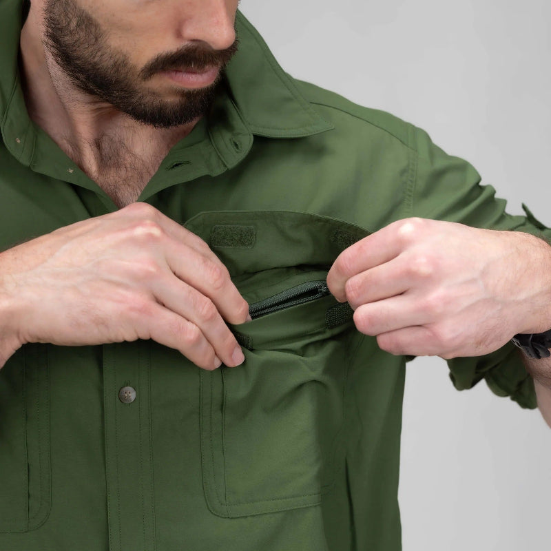 A close-up of a man wearing an olive green button-up shirt, demonstrating a zippered chest pocket by pulling the zipper tab with both hands.