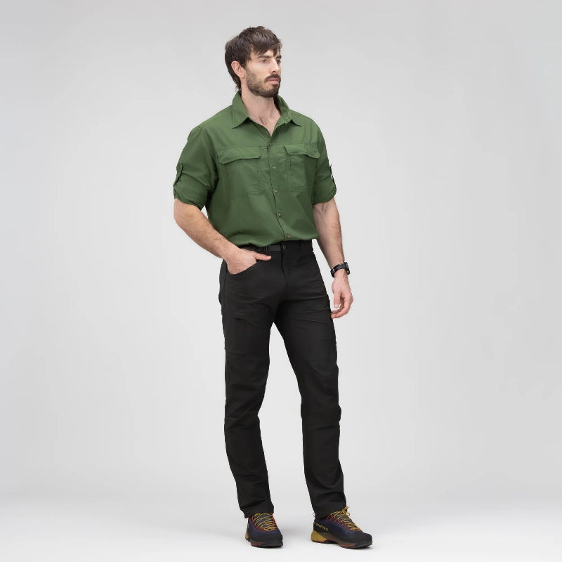 A man wearing an olive green long-sleeve button-up shirt and black pants, standing against a light gray studio background.