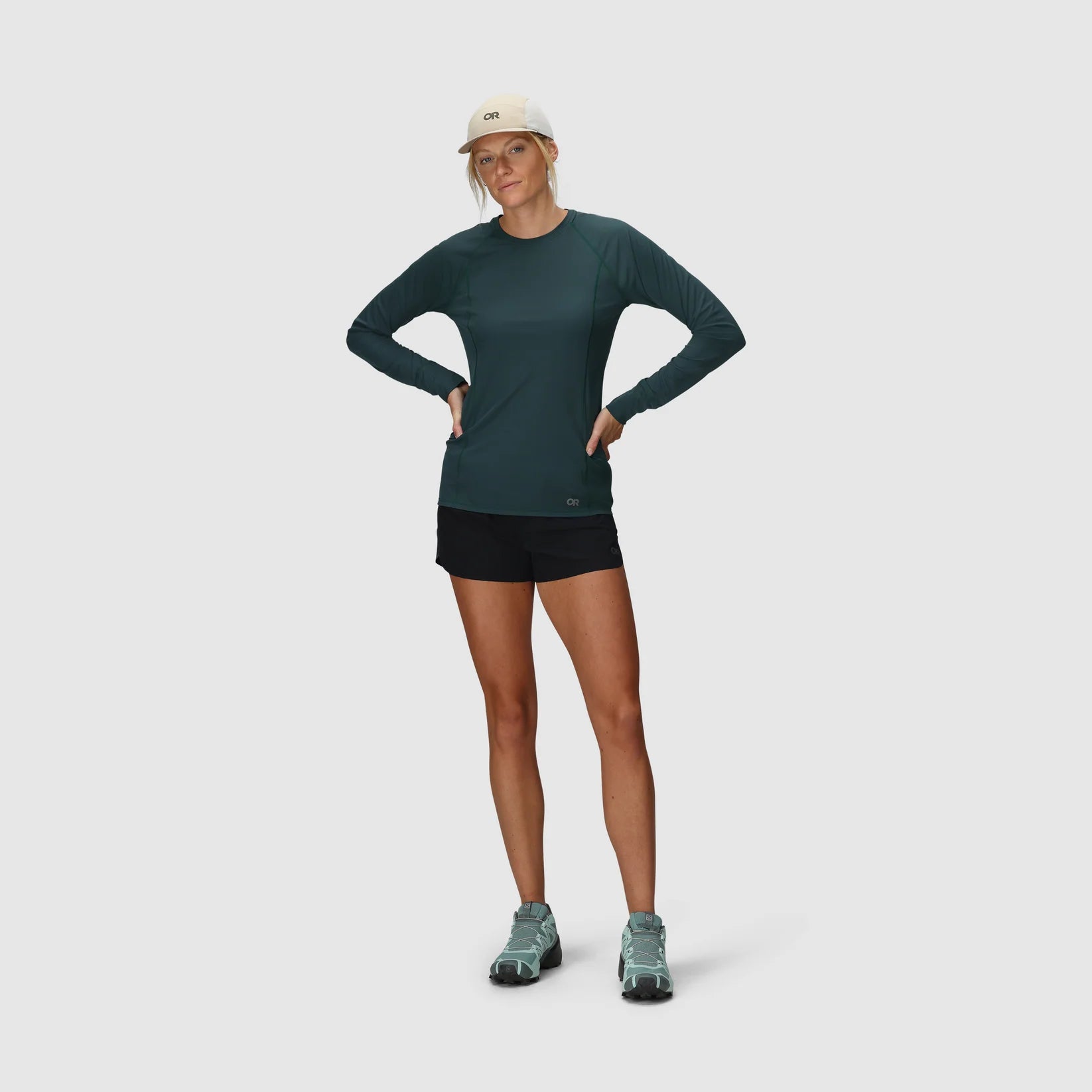 Woman wearing a green long-sleeve hiking top and black womens trekking shorts on a light gray background
