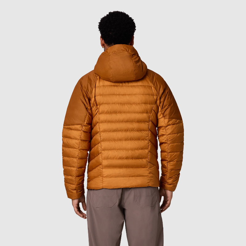 A man viewed from the back wearing a hooded puffer jacket in a warm camel-brown color, paired with taupe pants, against a light gray studio background.