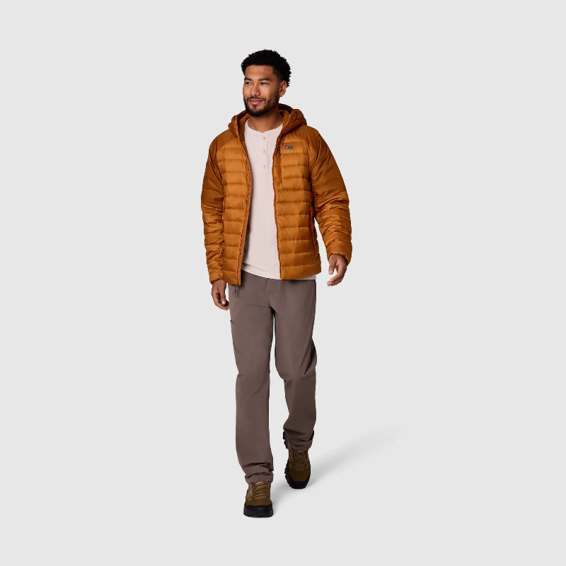 A man wearing a brown hooded puffer jacket over a light pink t-shirt, brown pants, and brown boots, walking against a light gray studio background.