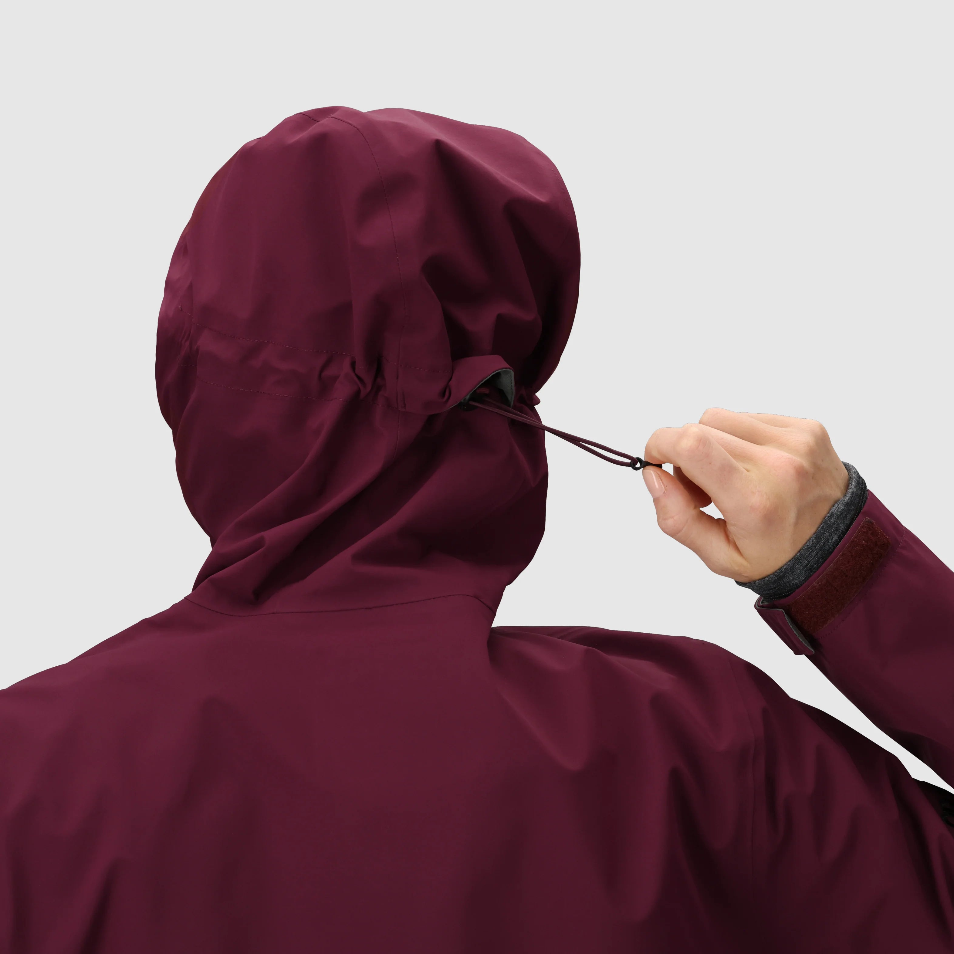 Person wearing a maroon hooded jacket adjusting the drawstring on a plain background