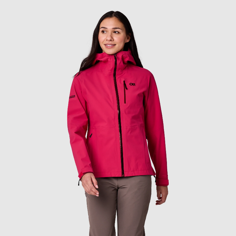 A woman modeling a bright pink Orvis women's waterproof jacket with black zippers and a black logo on the chest, paired with brown pants, against a light gray studio background.